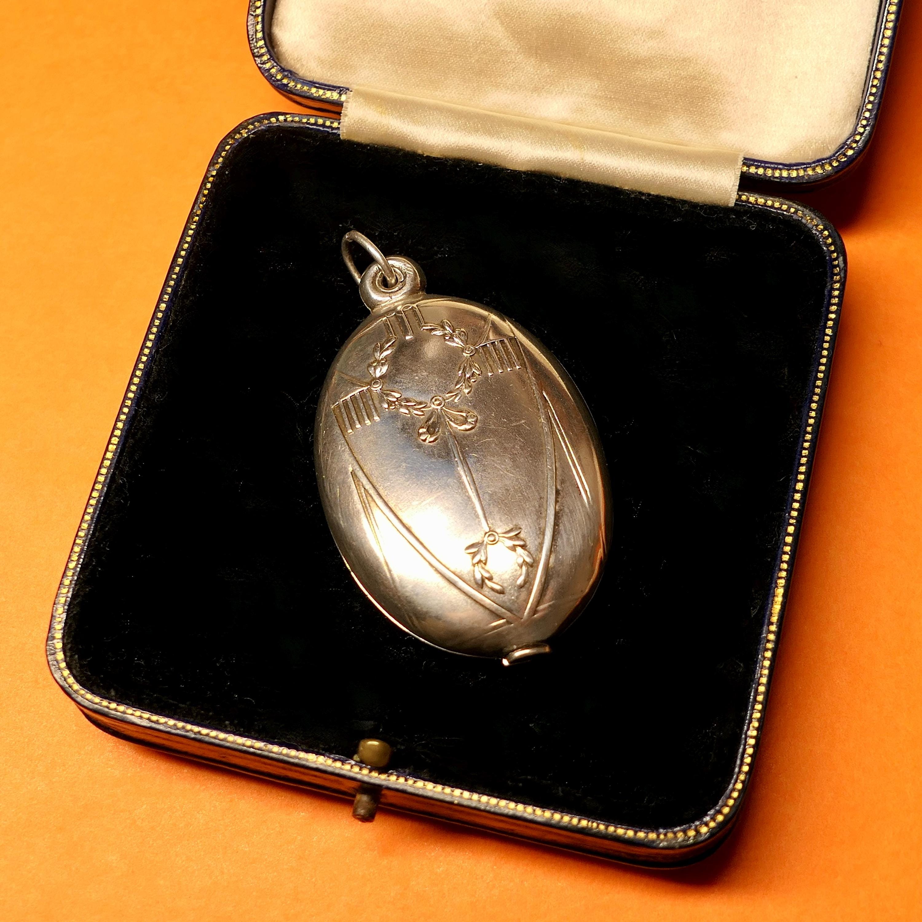 French antique sterling silver oval locket pendant with engraved wreath motifs resting in a velvet jewelry box